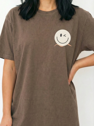 A close-up of a smiling woman in a Real Good Days Espresso brown 1717 heavyweight tee. The image highlights the premium garment-dyed cotton and the brand's winking smiley logo, accented by a signature apricot-colored wave that represents a calm, anti-hustle pace of life.