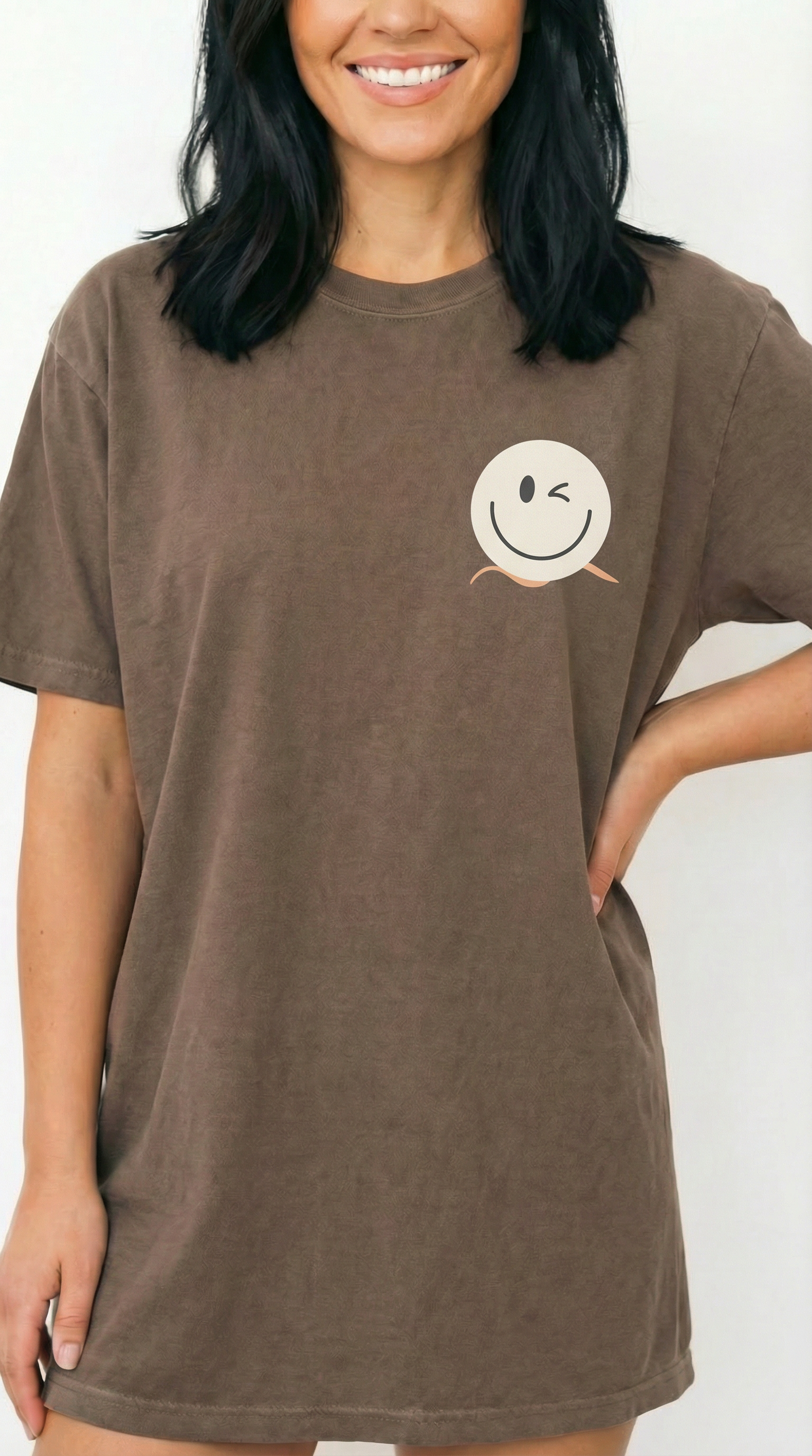 A close-up of a smiling woman in a Real Good Days Espresso brown 1717 heavyweight tee. The image highlights the premium garment-dyed cotton and the brand's winking smiley logo, accented by a signature apricot-colored wave that represents a calm, anti-hustle pace of life.